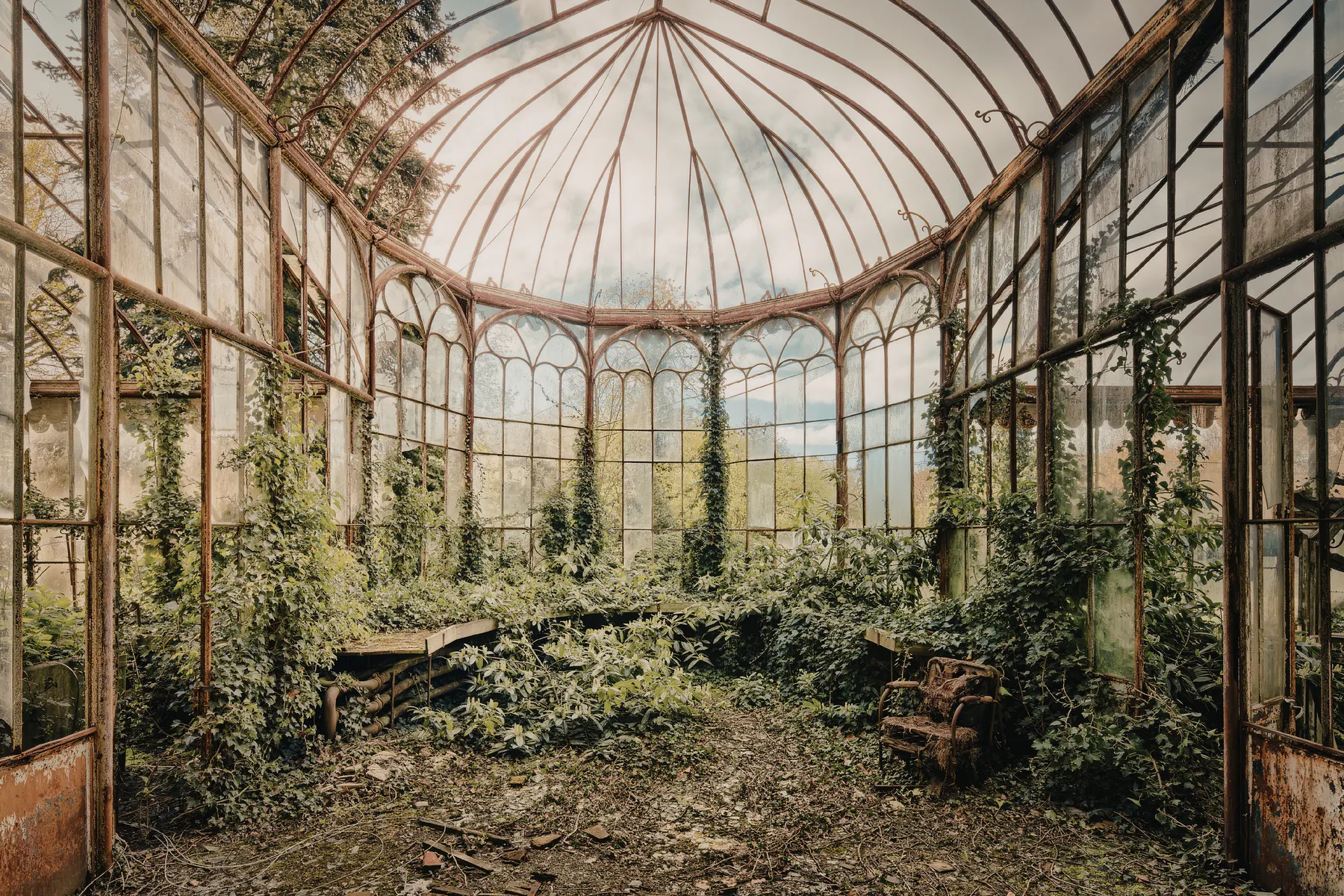 An ornate abandoned glasshouse with plants reclaiming the floor and climbing the ironwork